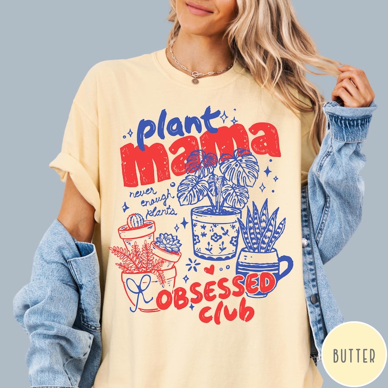 Puede incluir: Camiseta de color crema con un dise&ntilde;o gr&aacute;fico. El dise&ntilde;o incluye las palabras "plant mama" en rojo y azul, e ilustraciones de plantas en macetas. La frase "Obsessed Club" tambi&eacute;n est&aacute; en rojo. La camiseta tiene mangas cortas.
