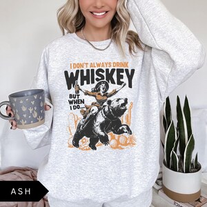 May include: A gray sweatshirt with a graphic print of a woman riding a bear. The text on the graphic reads "I don't always drink whiskey but when I do..."
