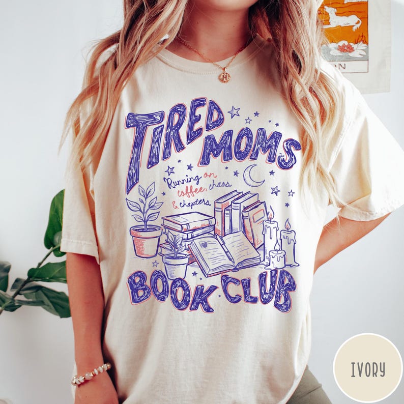 May include: Ivory-colored t-shirt with a graphic design. The design features the words "Tired Moms Book Club" in blue, along with illustrations of books, candles, plants, and stars. The text "Running on coffee, chaos & chapters" is also included.