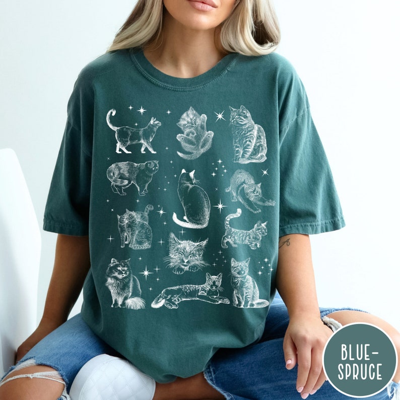 K&ouml;nnte beinhalten: Ein blaues T-Shirt mit einem wei&szlig;en Aufdruck von verschiedenen Katzen in verschiedenen Posen. Die Katzen sind von kleinen wei&szlig;en Sternen umgeben.