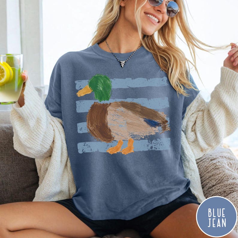 May include: A blue short-sleeve t-shirt featuring a graphic of a mallard duck with a green head, brown body, and orange feet. The duck is positioned over horizontal light blue distressed stripes. The shirt has a crew neckline and a relaxed fit.