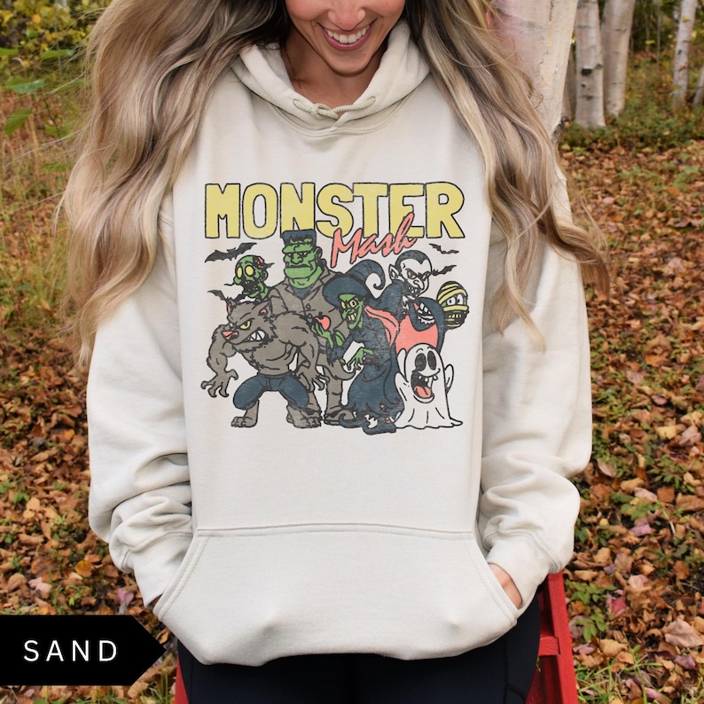 Puede incluir: Una sudadera con capucha beige claro con un estampado gr&aacute;fico de monstruos de dibujos animados. El texto "Monster Mash" est&aacute; impreso en amarillo y rojo en la sudadera.