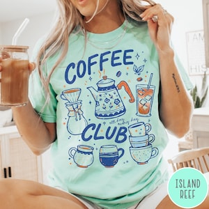 May include: A light green t-shirt with a "Coffee Club" graphic. The graphic includes a coffee pot, coffee cups, and the words "all day, every day". The person is holding a glass of iced coffee.