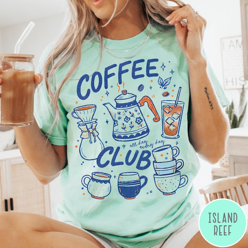 May include: A light green t-shirt with a "Coffee Club" graphic. The graphic includes a coffee pot, coffee cups, and the words "all day, every day". The person is holding a glass of iced coffee.