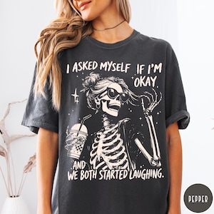 May include: Dark gray t-shirt featuring a skeleton graphic wearing sunglasses and holding a beverage. The text on the shirt reads, "I asked myself if I'm okay, and we both started laughing."