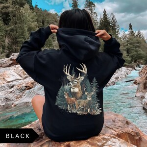 Op de afbeelding: Zwarte hoodie met een hertenafbeelding op de rug. De afbeelding toont een bok met geweien, twee herten en dennenbomen. Het woord "BLACK" staat onderaan de afbeelding.