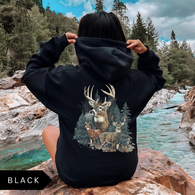 Op de afbeelding: Zwarte hoodie met een hertenafbeelding op de rug. De afbeelding toont een bok met geweien, twee herten en dennenbomen. Het woord "BLACK" staat onderaan de afbeelding.