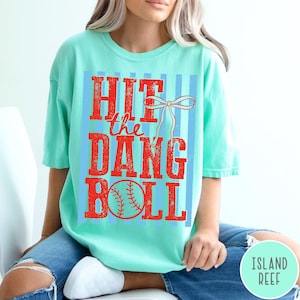 Pu&ograve; includere: T-shirt verde acqua chiaro con la scritta "HIT the DANG BALL" in lettere rosse. Il disegno include una palla da baseball e un fiocco. La maglietta &egrave; indossata con jeans blu.