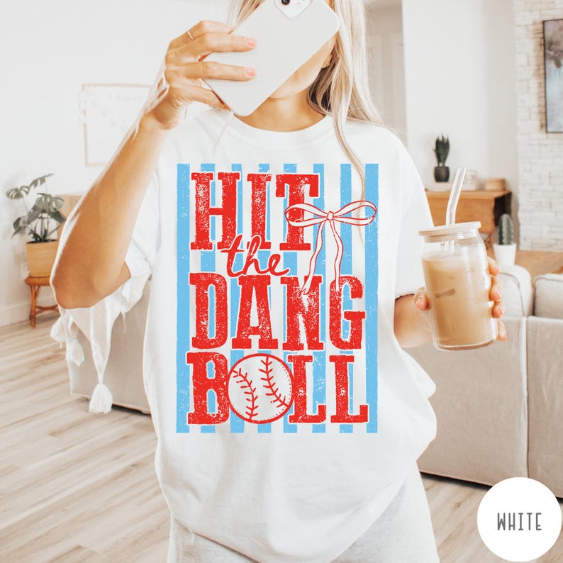 Pu&ograve; includere: T-shirt bianca a tema baseball. La maglietta presenta la scritta "HIT the DANG BALL" in rosso, con una grafica di baseball. Strisce verticali blu e un fiocco rosso accentuano il design.