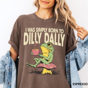 May include: Espresso-colored t-shirt featuring a whimsical frog illustration. The frog, with green skin and yellow accents, sits on a mushroom and holds a pink teacup. The text "I WAS SIMPLY BORN TO DILLY DALLY" is printed above the frog.