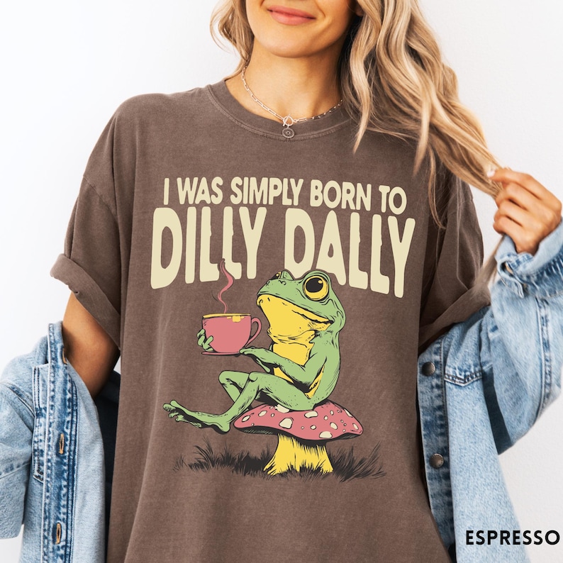 May include: Espresso-colored t-shirt featuring a whimsical frog illustration. The frog, with green skin and yellow accents, sits on a mushroom and holds a pink teacup. The text "I WAS SIMPLY BORN TO DILLY DALLY" is printed above the frog.