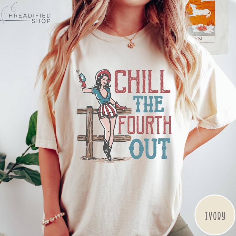 May include: Ivory-colored t-shirt with a graphic of a woman in patriotic attire holding a firecracker, leaning on a wooden fence. The text "CHILL THE FOURTH OUT" is printed in red and blue.