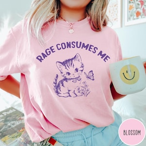 Puede incluir: Una camiseta rosa con un gr&aacute;fico morado de un gato con una mariposa y flores. El texto "Rage Consumes Me" est&aacute; impreso en la camiseta.