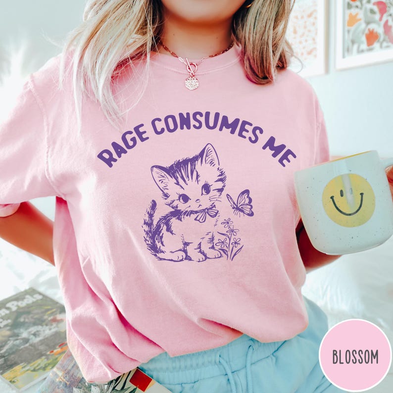 Puede incluir: Una camiseta rosa con un gr&aacute;fico morado de un gato con una mariposa y flores. El texto "Rage Consumes Me" est&aacute; impreso en la camiseta.