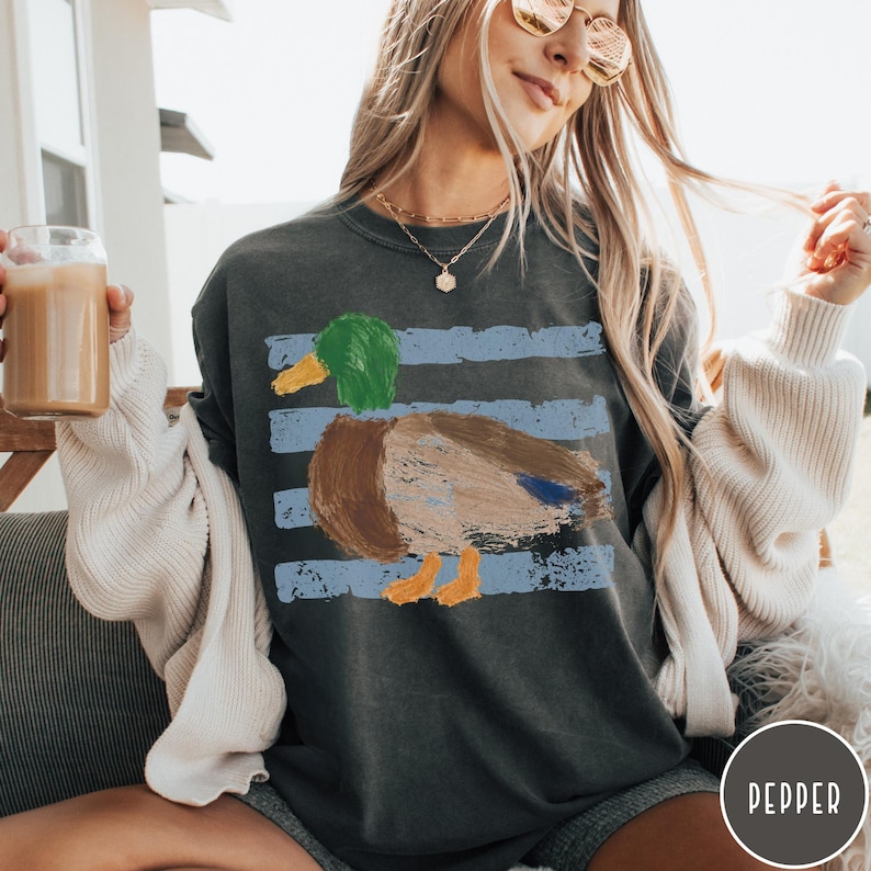 May include: A charcoal gray crewneck sweatshirt featuring a painterly graphic of a mallard duck in shades of green, brown, and blue, set against horizontal distressed light blue stripes. The design is centered on the chest of the garment.