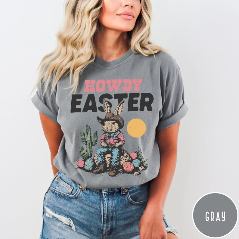 Puede incluir: Camiseta gris con el texto "HOWDY EASTER" en rosa y negro. El gr&aacute;fico presenta un conejo con sombrero de vaquero sentado en una roca con huevos de Pascua y un cactus. Un c&iacute;rculo amarillo est&aacute; a la derecha del conejo.