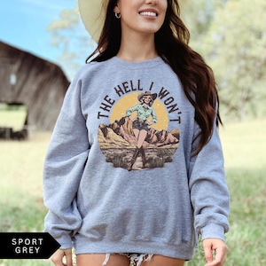 May include: A gray sweatshirt with the text "The Hell I Won't" in a vintage style font. The text is surrounded by a graphic of a woman in a cowboy hat and boots standing in a desert landscape.