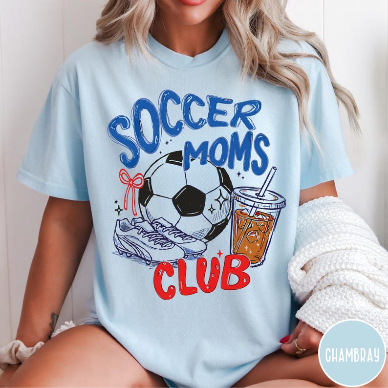 May include: Light blue t-shirt with a graphic that reads "Soccer Moms Club." The design includes a soccer ball, cleats, a drink, and a red bow. The shirt is a comfortable, casual style.