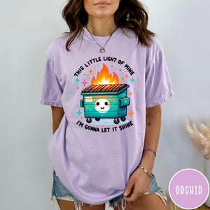May include: A purple t-shirt with a graphic of a green dumpster on fire with the text "This little light of mine, I'm gonna let it shine".