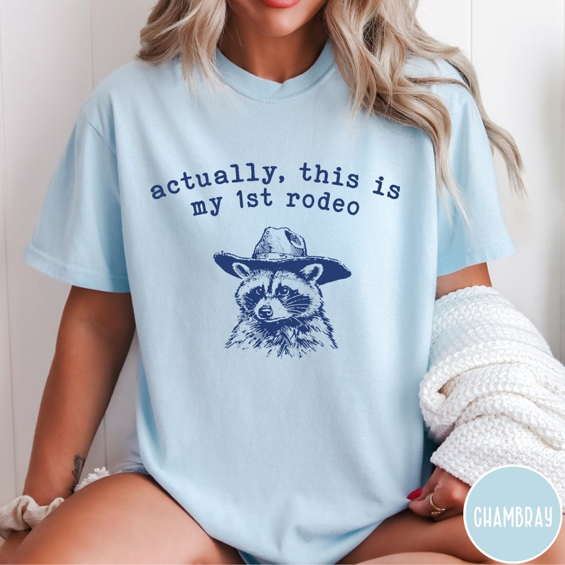 May include: Light blue t-shirt with a raccoon wearing a cowboy hat and the text "actually, this is my 1st rodeo".