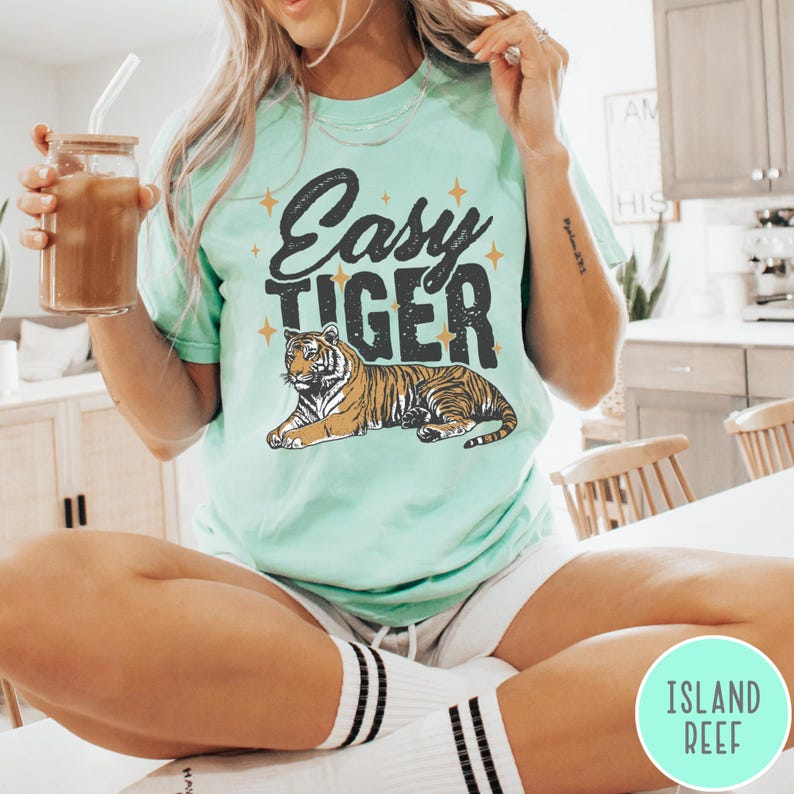 May include: A light green t-shirt with the text "Easy Tiger" in black lettering and a stylised orange tiger illustration. The shirt features a relaxed fit and a crew neck.