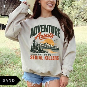 May include: A sand-colored crewneck sweatshirt featuring a graphic print with the text ADVENTURE Awaits BUT SO DO SERIAL KILLERS. The design includes a dark green mountain range, a yellow sun, a lake, pine trees, and small black birds.