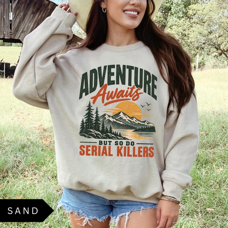 May include: A sand-colored crewneck sweatshirt featuring a graphic print with the text ADVENTURE Awaits BUT SO DO SERIAL KILLERS. The design includes a dark green mountain range, a yellow sun, a lake, pine trees, and small black birds.