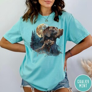 May include: A light blue t-shirt featuring a watercolor design of two brown dogs with a full moon and forest background. The shirt has short sleeves and a relaxed fit. The text "CHALKY MINT" is in a circle on the bottom right.