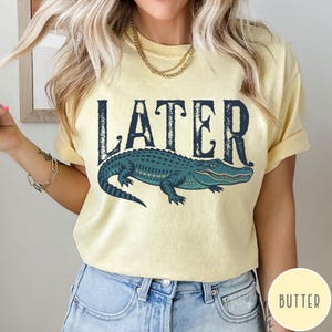 May include: A light yellow t-shirt with the word "LATER" in dark blue block letters above a teal alligator illustration. The alligator is detailed with lighter green and blue accents.