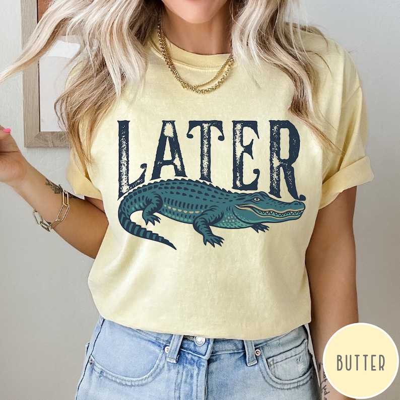 May include: A light yellow t-shirt with the word "LATER" in dark blue block letters above a teal alligator illustration. The alligator is detailed with lighter green and blue accents.