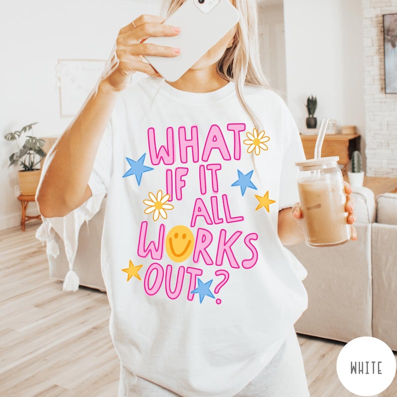 May include: A white short-sleeve t-shirt featuring the pink text "WHAT IF IT ALL WORKS OUT?" printed on the front. The design includes yellow smiley faces, yellow daisies, and blue and yellow stars scattered around the lettering.