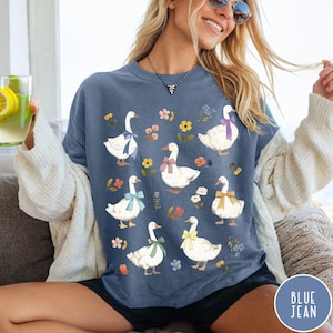 K&ouml;nnte beinhalten: Ein T-Shirt in Jeansblau mit wei&szlig;em Hintergrund und einem Muster aus wei&szlig;en Enten mit bunten Schleifen und floralen Akzenten.