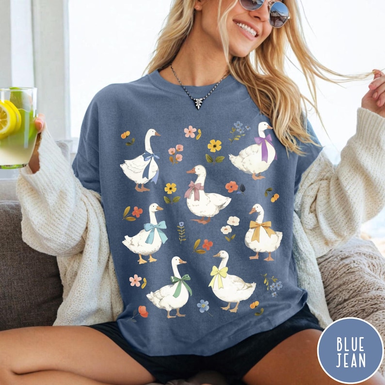 K&ouml;nnte beinhalten: Ein T-Shirt in Jeansblau mit wei&szlig;em Hintergrund und einem Muster aus wei&szlig;en Enten mit bunten Schleifen und floralen Akzenten.