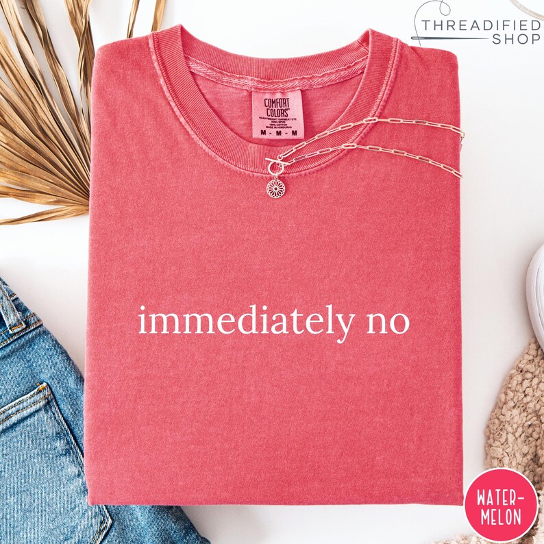 May include: A coral-colored t-shirt with the words "immediately no" printed in white. A delicate silver necklace with a sunburst pendant rests on the shirt. The shirt is folded neatly, with a tag visible.