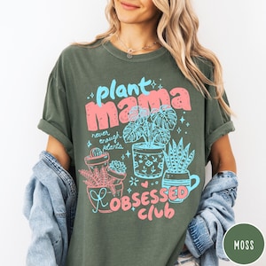 Puede incluir: Camiseta verde musgo con la frase "plant mama" en rosa y azul, junto con ilustraciones de plantas en macetas. La camiseta tambi&eacute;n presenta las palabras "never enough plants" y "obsessed club".
