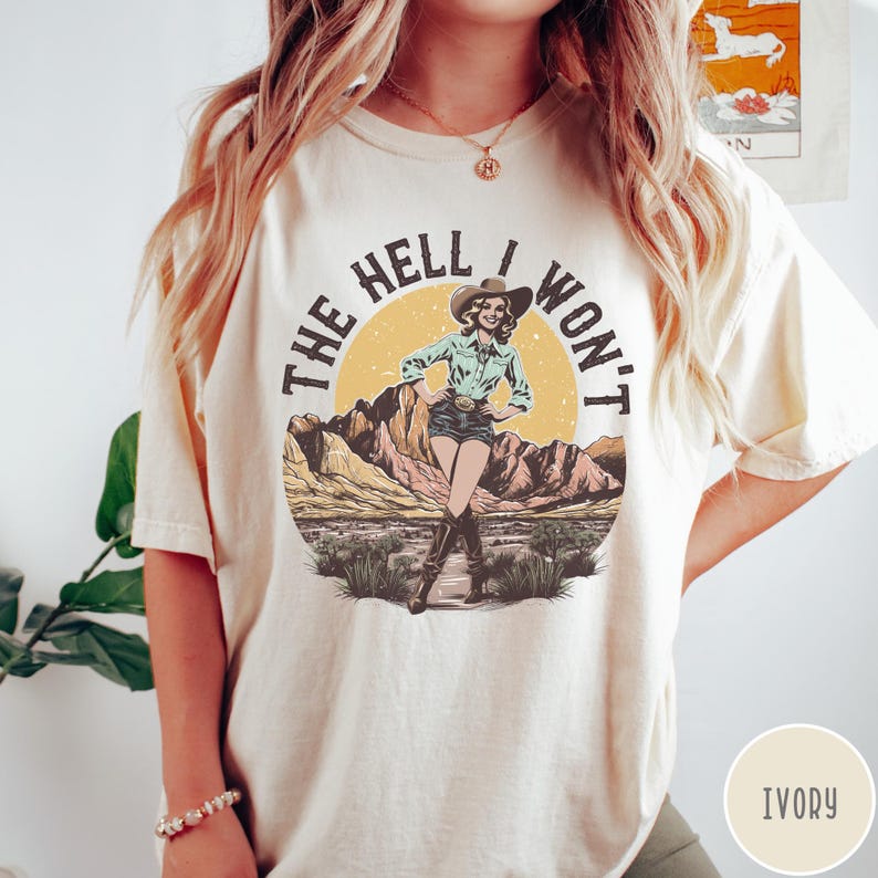 Może przedstawiać: Biały t-shirt z grafiką w stylu vintage przedstawiającą kobietę w kowbojskim kapeluszu i dżinsach. Grafika głosi "The Hell I Won't". Koszulka dostępna jest w kolorze kości słoniowej.