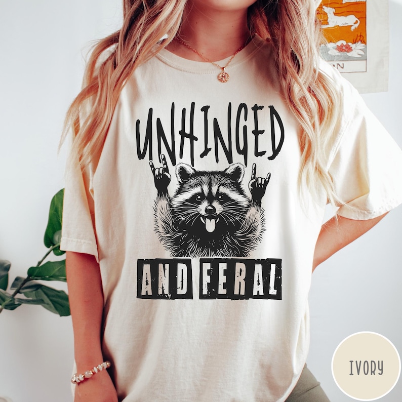 May include: A white t-shirt with a black graphic of a raccoon with the text "Unhinged and Feral". The raccoon is wearing a rock and roll hand gesture. The shirt is available in ivory.
