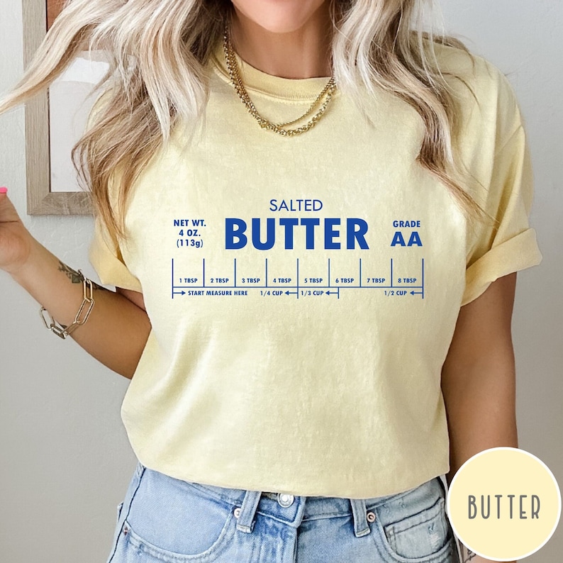 May include: A yellow t-shirt with a blue graphic of a stick of butter with the text "SALTED BUTTER GRADE AA" and a measurement guide for tablespoons and cups. The text "BUTTER" is printed in a smaller font at the bottom of the shirt.