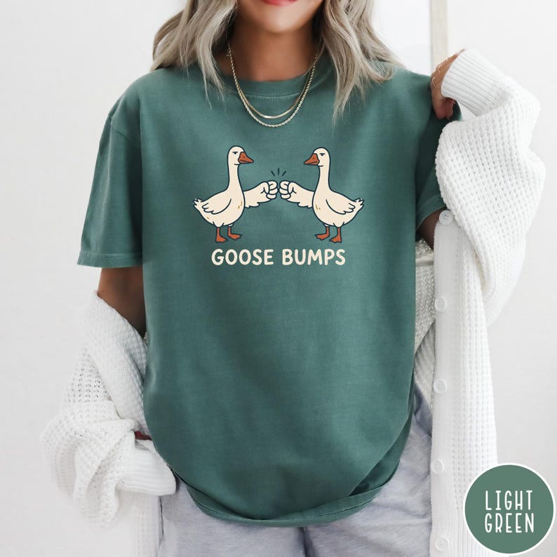 Puede incluir: Una camiseta de manga corta de color verde oscuro con un gr&aacute;fico de dos gansos blancos chocando los pu&ntilde;os. Debajo de los gansos, el texto GOOSE BUMPS est&aacute; impreso en una fuente de color crema. La camiseta tiene cuello redondo y corte casual.
