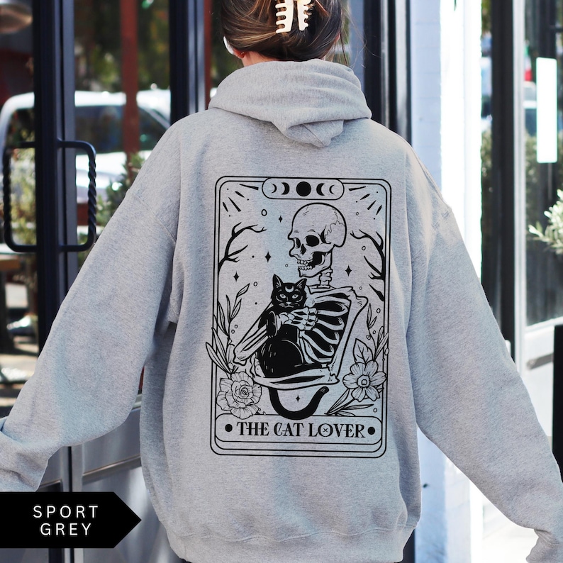 May include: A gray hoodie with a black and white illustration of a skeleton holding a black cat. The illustration is in the shape of a tarot card and says "The Cat Lover" at the bottom.
