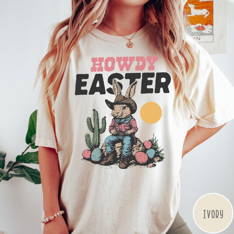 Puede incluir: Camiseta de color marfil con las palabras "HOWDY EASTER" en rosa y negro. El gr&aacute;fico presenta un conejo vaquero sentado en una roca con un cactus, huevos de Pascua y un sol amarillo. La camiseta es de corte holgado.