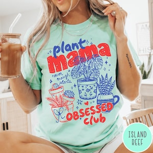 Puede incluir: Camiseta verde claro con las palabras "plant mama" en rojo y azul, junto con ilustraciones de plantas en macetas. La camiseta tambi&eacute;n presenta el texto "never enough plants" y "obsessed club".