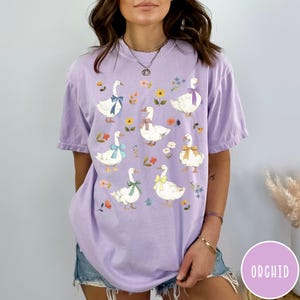 K&ouml;nnte beinhalten: Ein lila T-Shirt mit einem Blumenmuster und einem Design aus wei&szlig;en G&auml;nsen mit bunten Schleifen. Die G&auml;nse sind symmetrisch auf dem Shirt angeordnet.