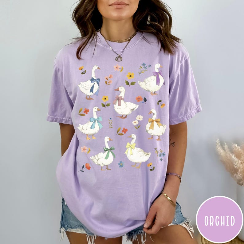 K&ouml;nnte beinhalten: Ein lila T-Shirt mit einem Blumenmuster und einem Design aus wei&szlig;en G&auml;nsen mit bunten Schleifen. Die G&auml;nse sind symmetrisch auf dem Shirt angeordnet.