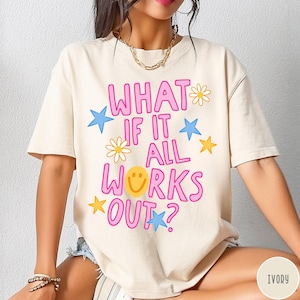 May include: An ivory short-sleeved t-shirt featuring the pink text "WHAT IF IT ALL WORKS OUT?" with a yellow smiley face replacing the letter O. The design includes scattered yellow daisies and blue and yellow stars printed on the front.