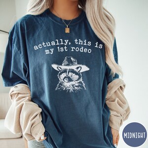 May include: A dark blue t-shirt with the text "actually, this is my 1st rodeo" and a black and white illustration of a raccoon wearing a cowboy hat.