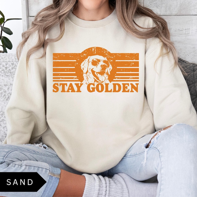 Puede incluir: Una sudadera de color crema con un gr&aacute;fico de estilo vintage de un golden retriever con gafas de sol. El gr&aacute;fico es de color naranja y dice "Stay Golden".