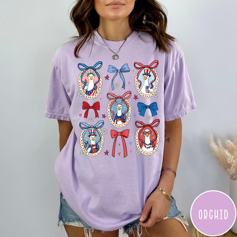 May include: A light purple t-shirt featuring a whimsical design of cartoon geese in oval frames, each adorned with patriotic bows and accessories. The geese are dressed in red, white, and blue, with floral accents. The shirt has a relaxed fit.