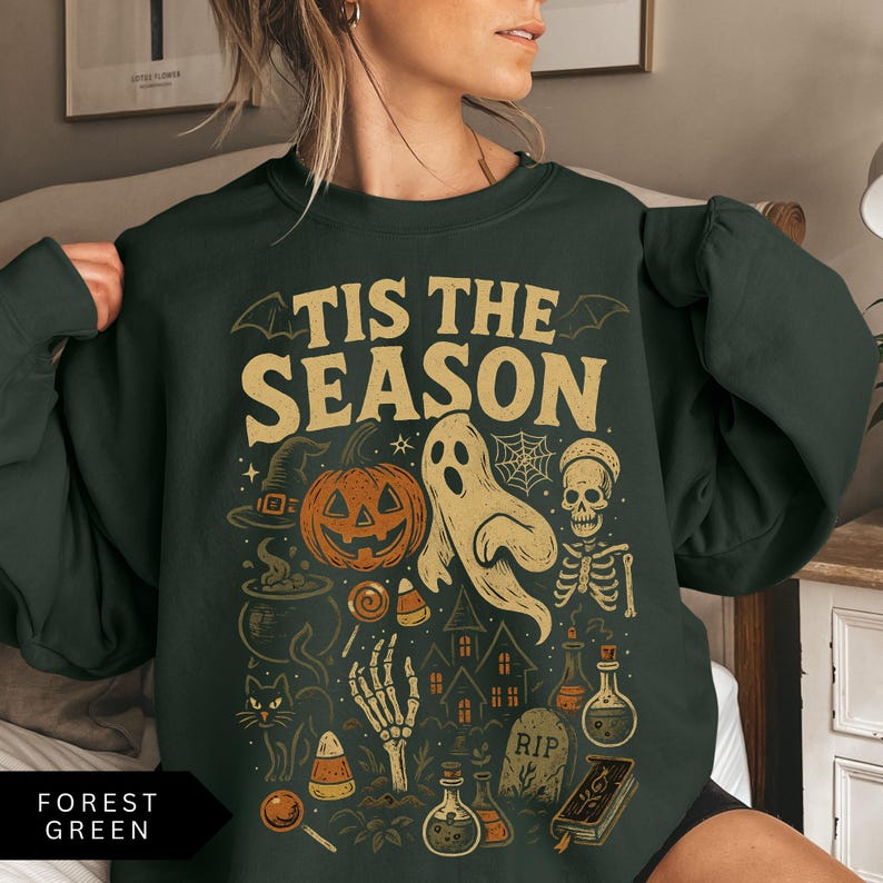 Puede incluir: Sudadera verde bosque con un dise&ntilde;o de Halloween. El dise&ntilde;o incluye una calabaza, un fantasma, una calavera y el texto "TIS THE SEASON" en un estilo vintage. Elementos adicionales incluyen un sombrero de bruja, una telara&ntilde;a y botellas de pociones.
