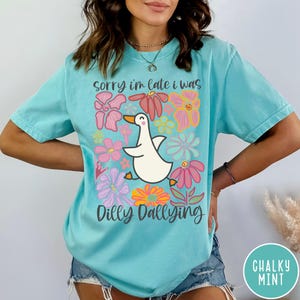 May include: A turquoise blue t-shirt with a white duck and colorful flowers. The text on the shirt reads "Sorry I'm late I was dilly dallying".